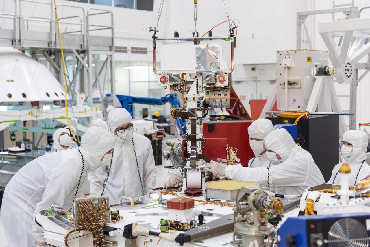 Engineers in white cleanroom suits and masks carefully fitting a component onto the Mars Perseverance rover
