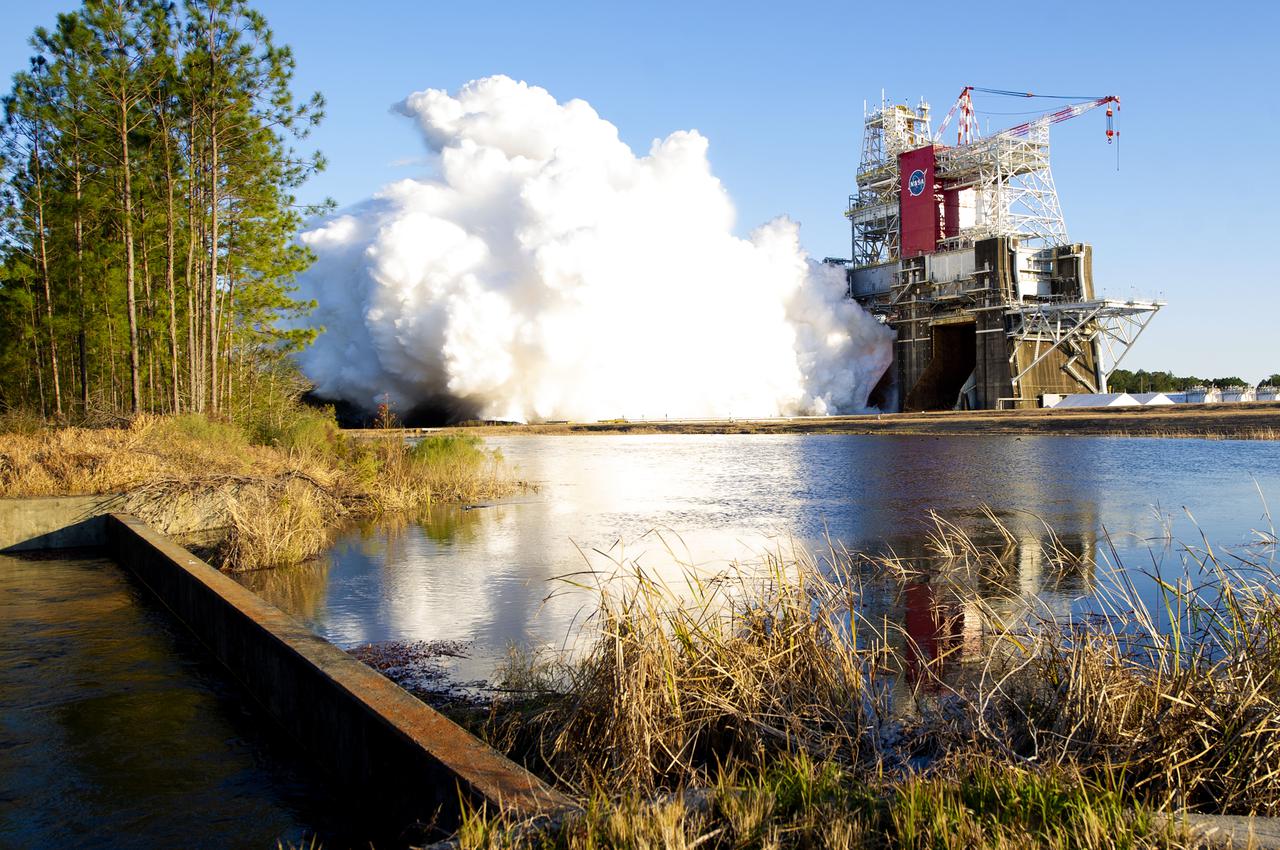 A rocket engine firing on a test stand beside a lake, a huge white exhaust plume rolling sideways across the water