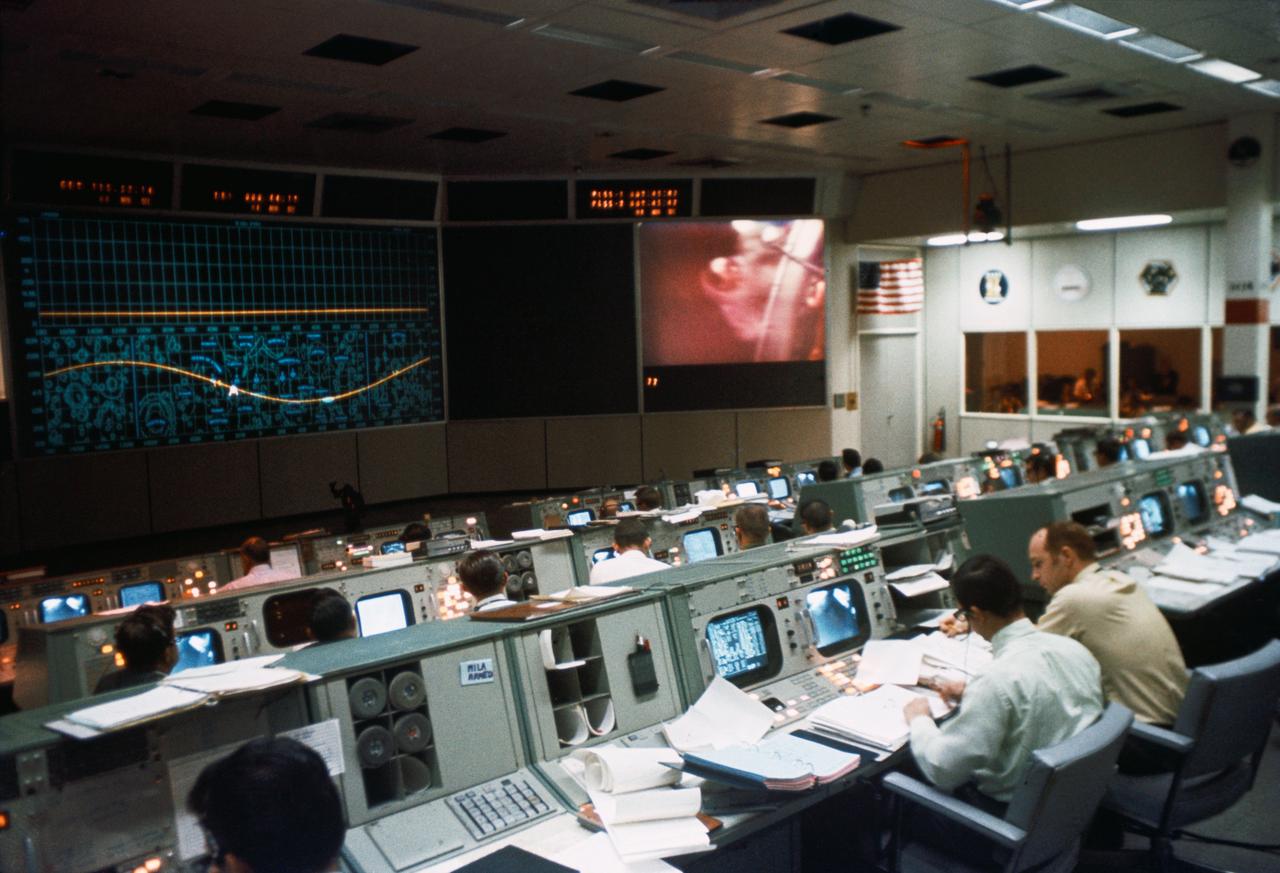 Flight controllers at consoles facing a large screen showing a plotted trajectory curve