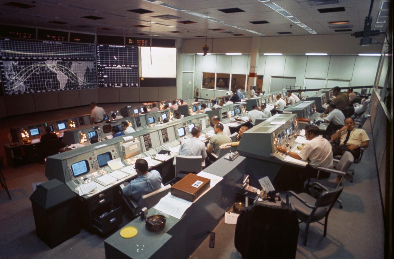 Rows of flight controllers at consoles in NASA's Mission Operations Control Room during Apollo 7, with a world map on the front screen