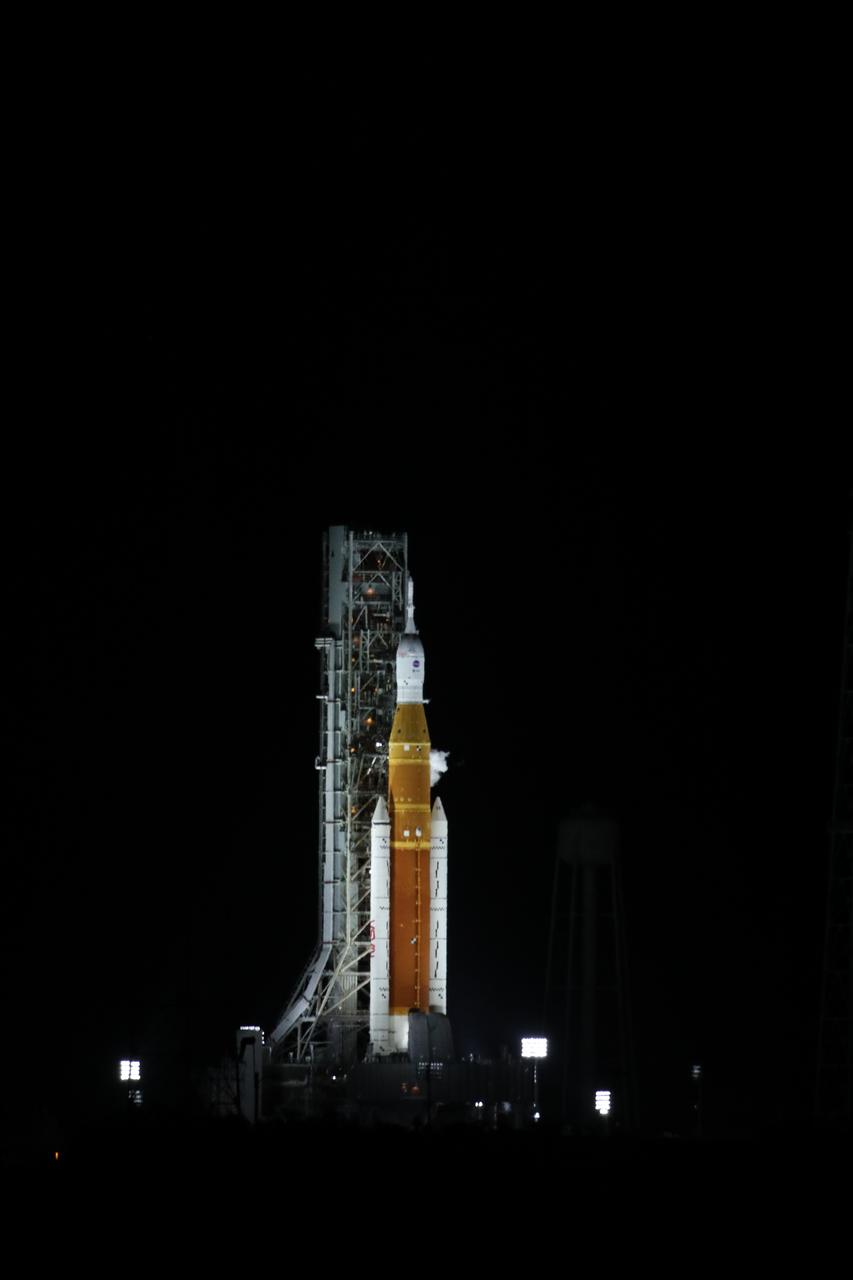 A rocket lit by floodlights on a launch pad against a completely black night sky