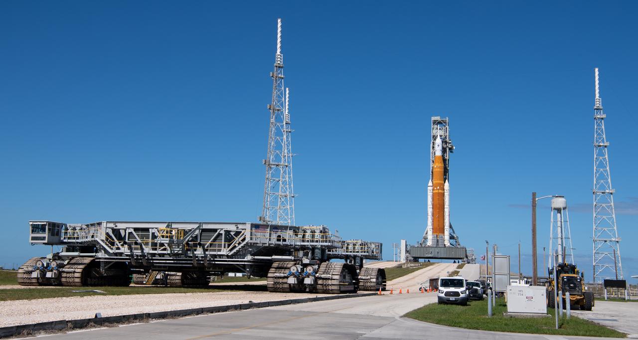 A large orange and white rocket standing on a distant launch pad, with the tracked crawler-transporter parked in the foreground