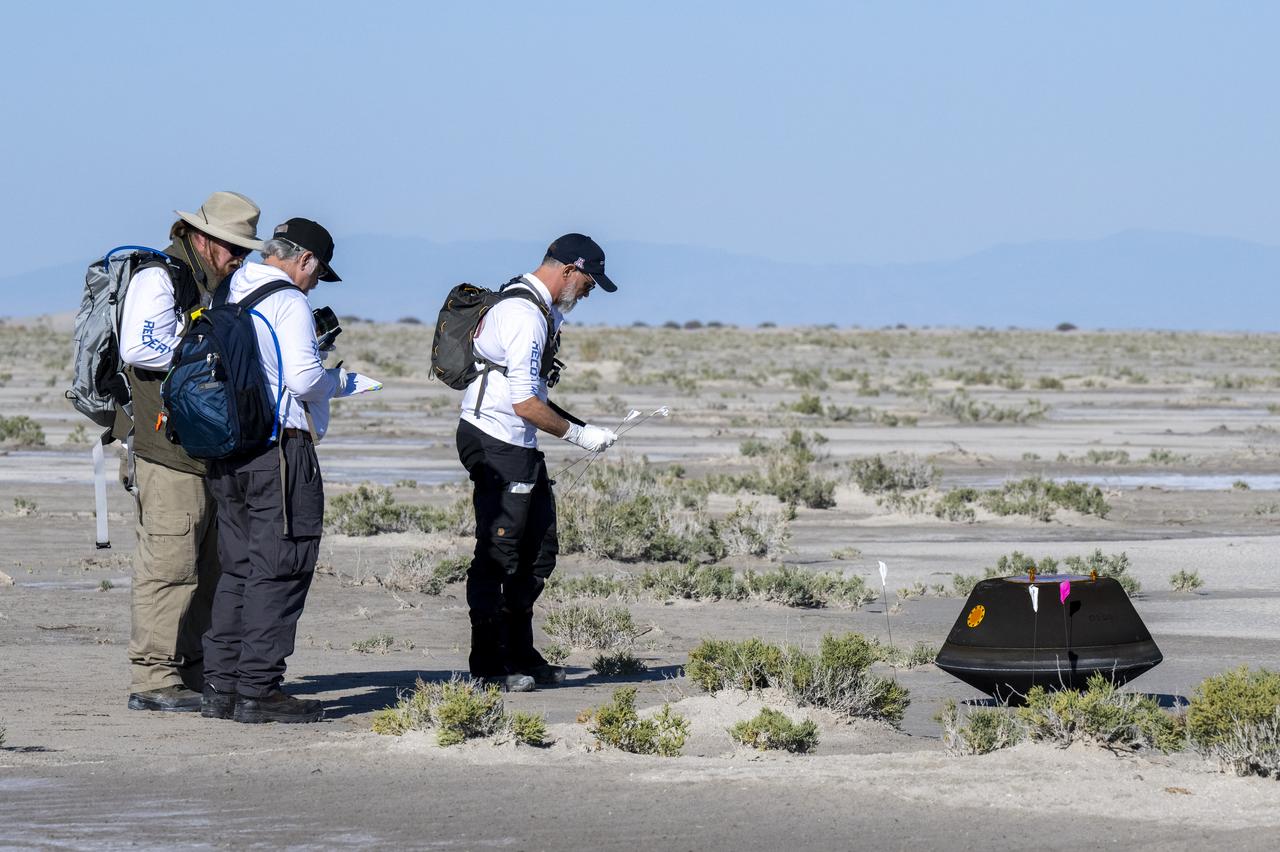 Three recovery team members standing on desert scrubland beside a small dark cone-shaped capsule on the ground