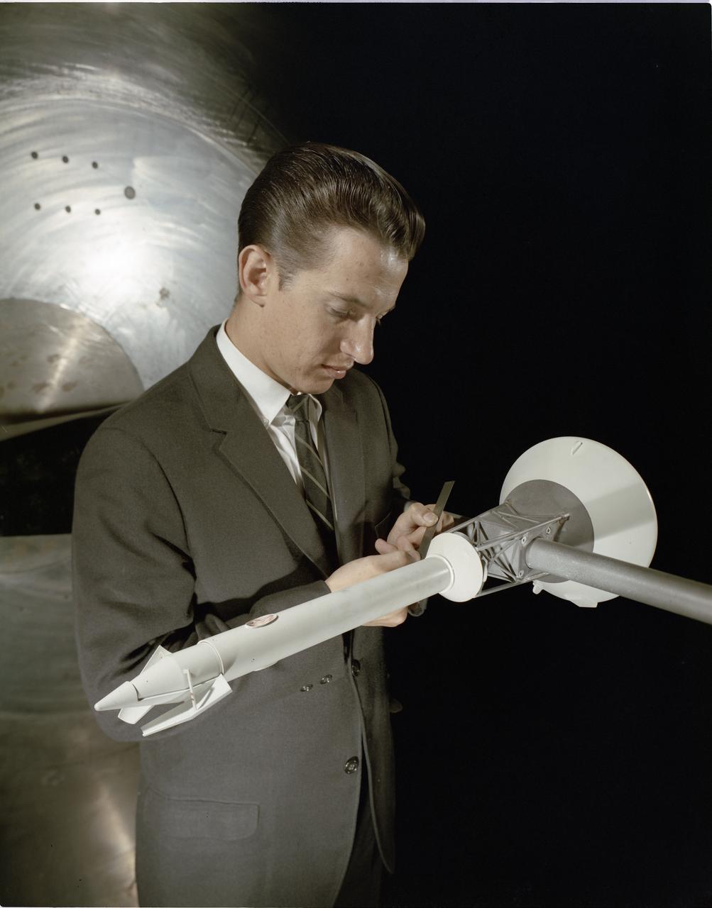 An engineer in a suit holding a small model rocket mounted on a sting in a wind tunnel, measuring it with a rule