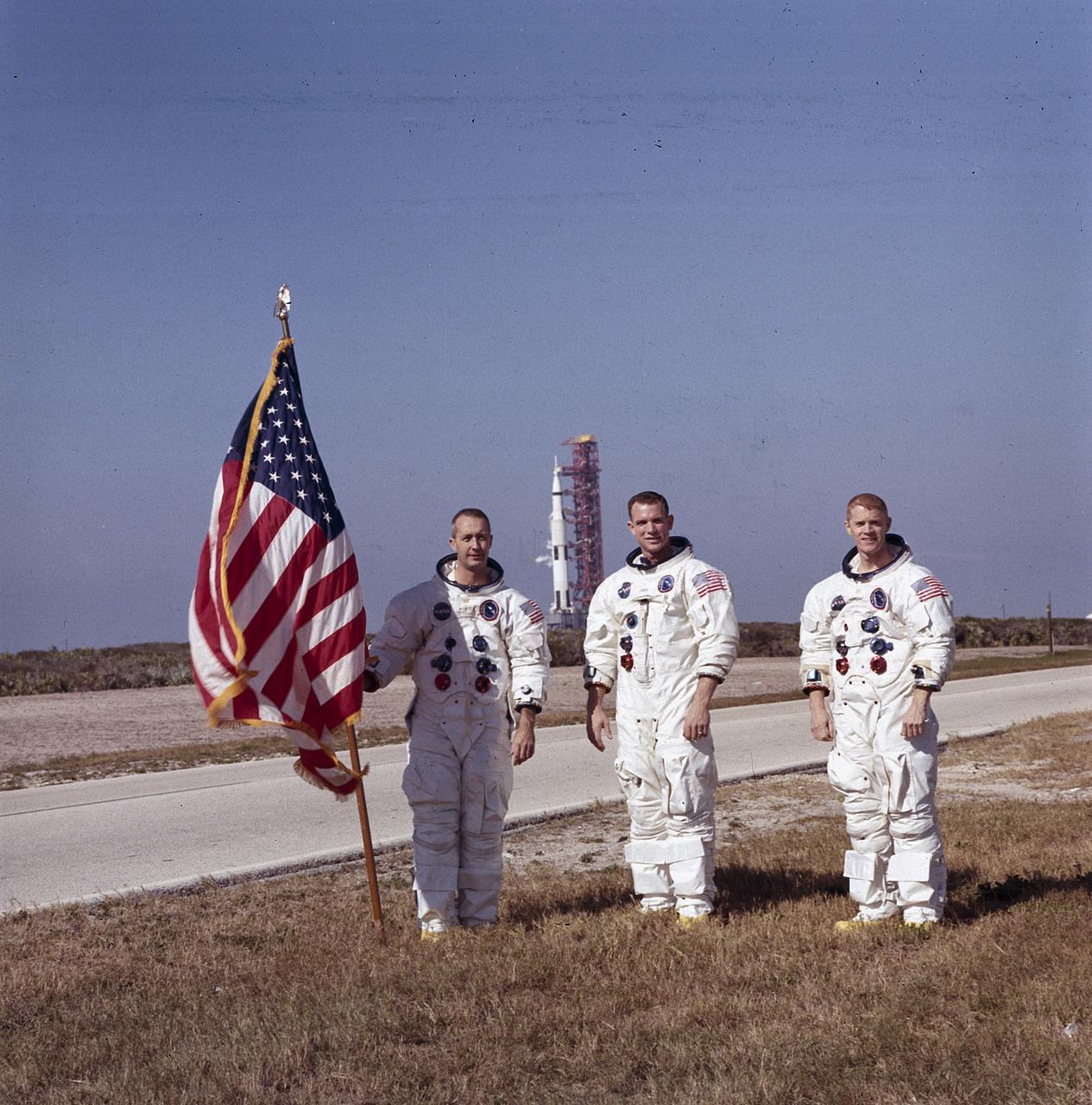 Three Apollo astronauts in white pressure suits standing in front of an American flag, with a Saturn V rocket on its pad in the distance